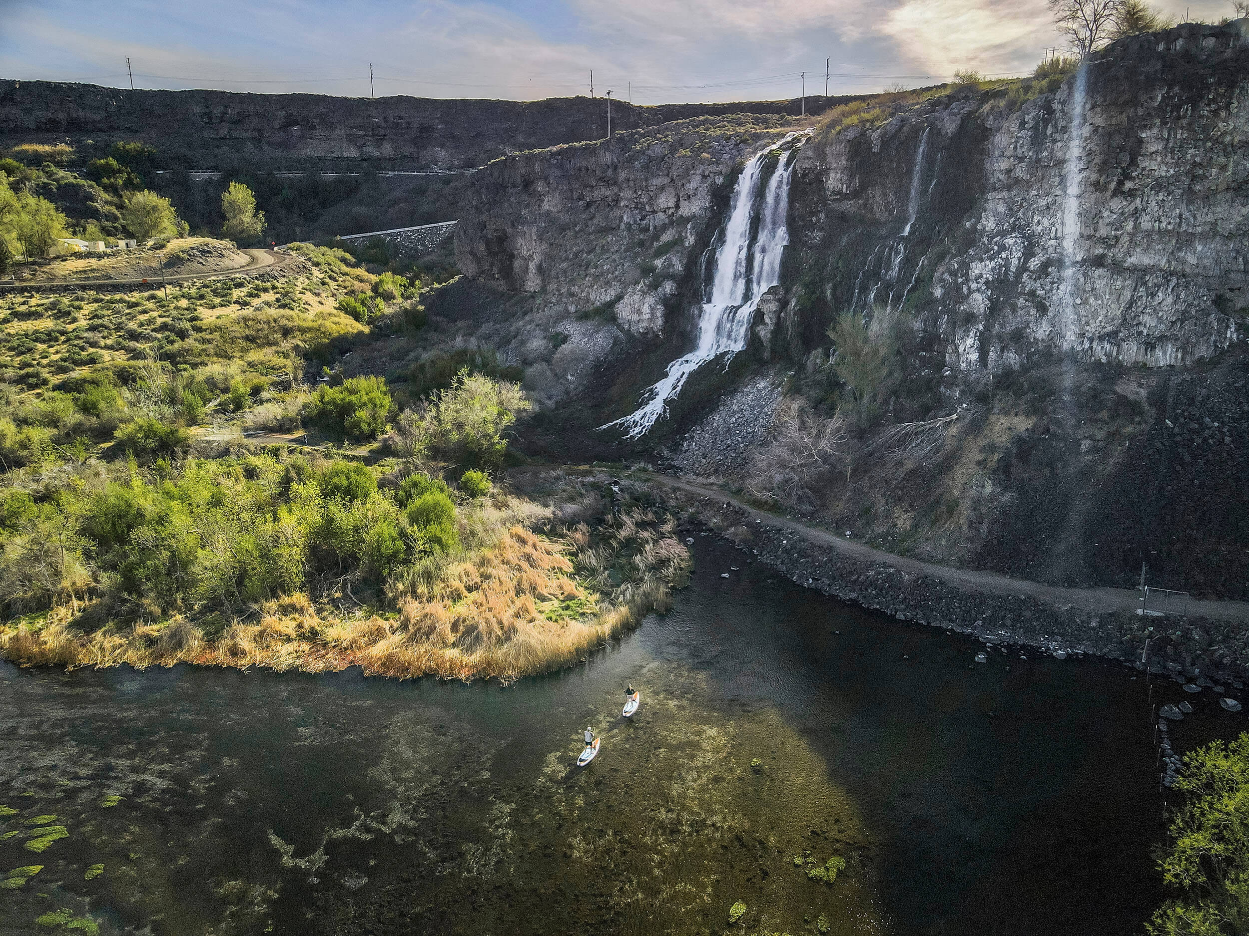 A bird's-eye view of two people paddle boarding on white paddle boards below a waterfall.