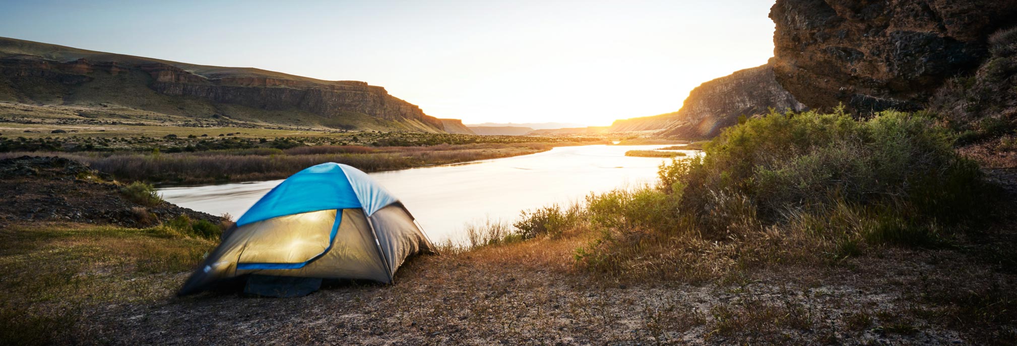 A scenic shot of a blue and white tent up set up in front of a lake in a canyon at sunrise.