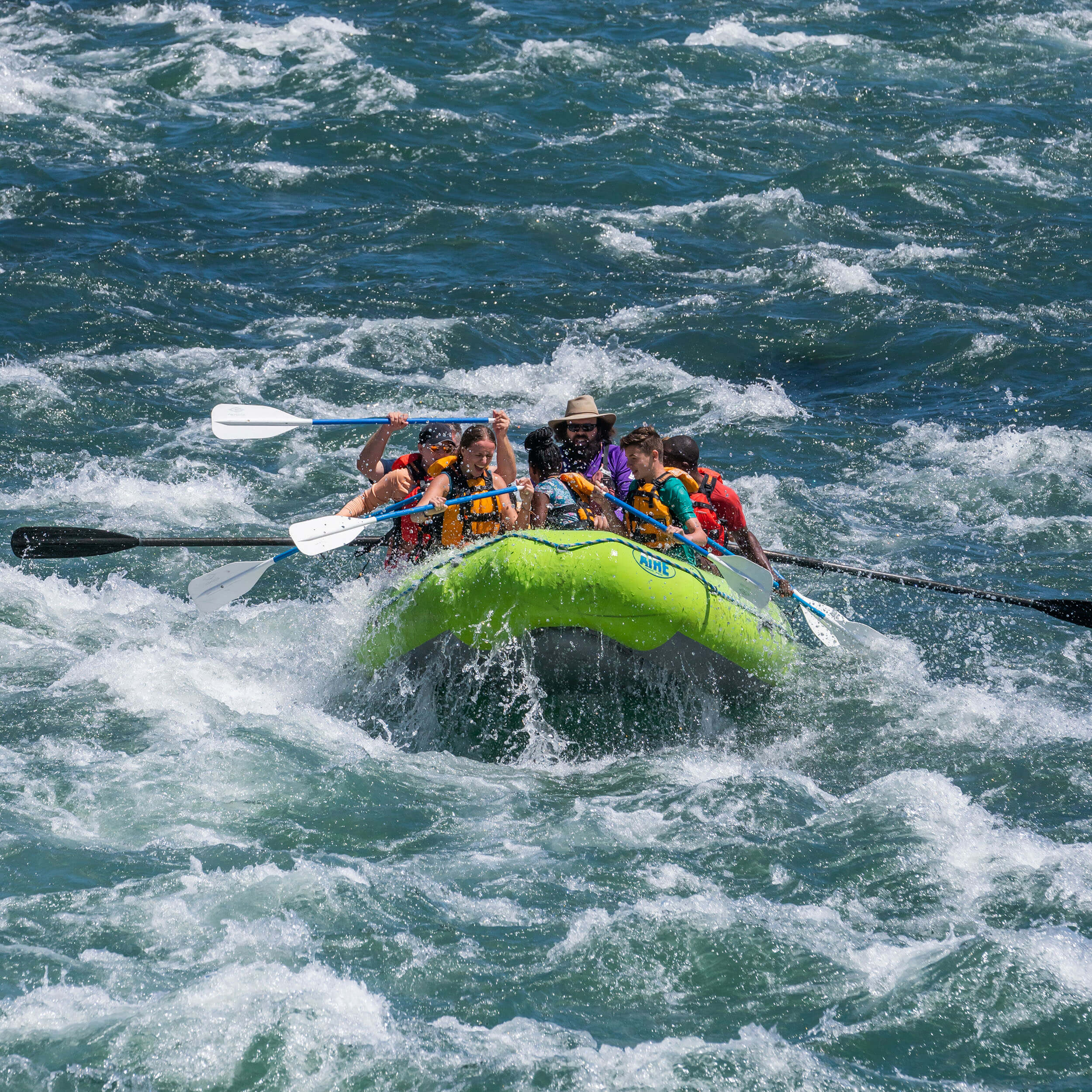 A group of children and adults in life jackets white water rafting with a guide in a neon green raft.